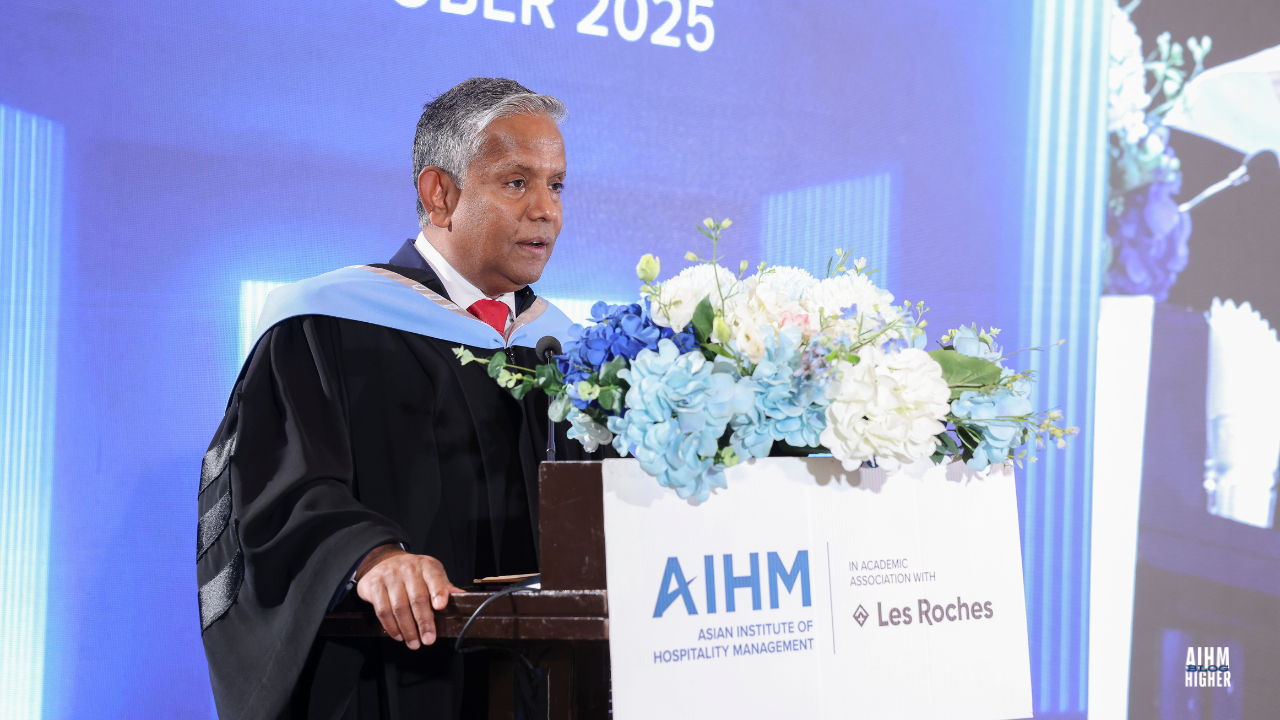 Mr. Dillip Rajakarier, Group CEO of Minor International and CEO of Minor Hotels in academic dress speaks at the podium during the AIHM Graduation Ceremony 2025. The stage backdrop highlights the event title and date in white text on a blue background.