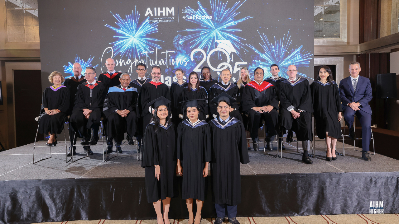 AIHM graduates BBA Cohort 2 stand at the front of the stage with faculty members behind them, marking their proud achievement during the Graduation Ceremony 2025. The background shows the message “Congratulations Class of 2025” with bright blue fireworks.