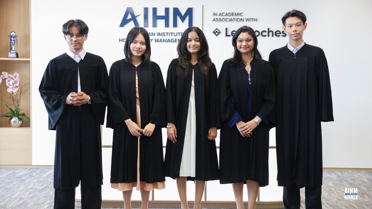 Five BTEC L3 graduates stand together in academic gowns, smiling proudly in front of the AIHM and Les Roches logo wall. The setting reflects a professional and celebratory atmosphere before the AIHM Graduation Ceremony 2025.