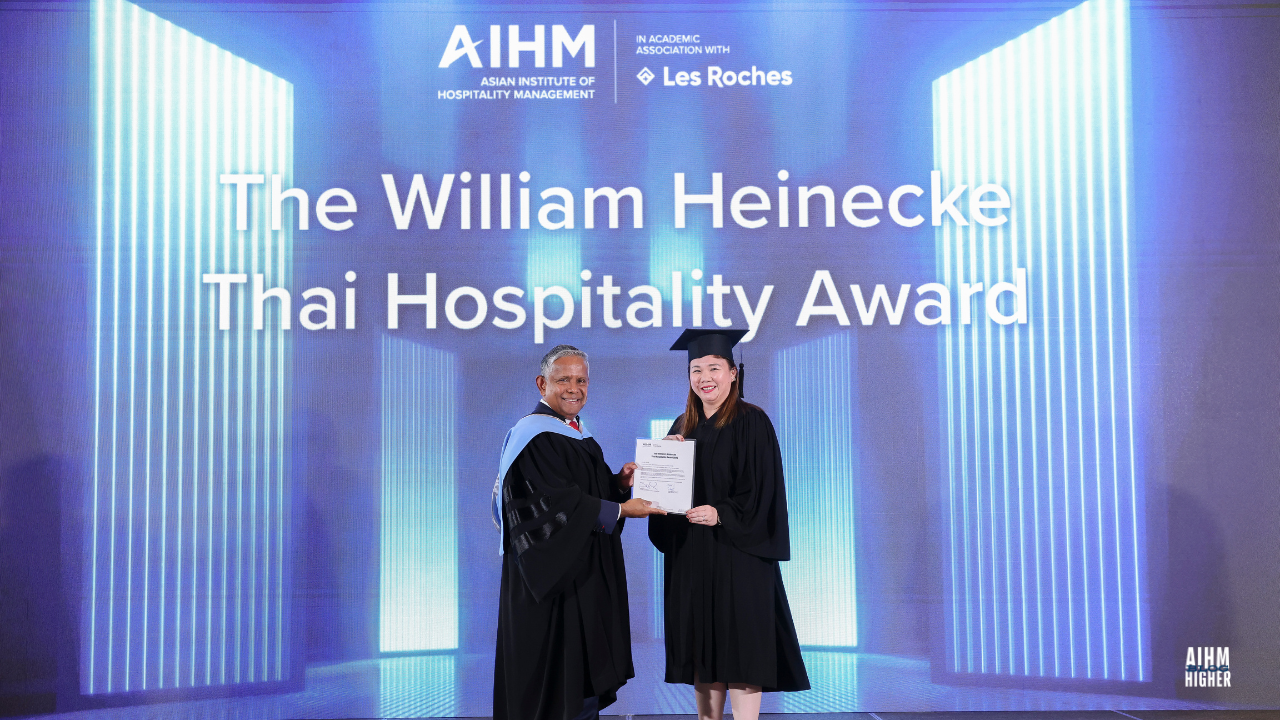 A Rashelle Capanas Gomez CIHA3 smiles as she accepts The William Heinecke Thai Hospitality Award from a Mr. Dillip Rajakarier on stage. The background displays the AIHM and Les Roches logos and the award title in bright white text.