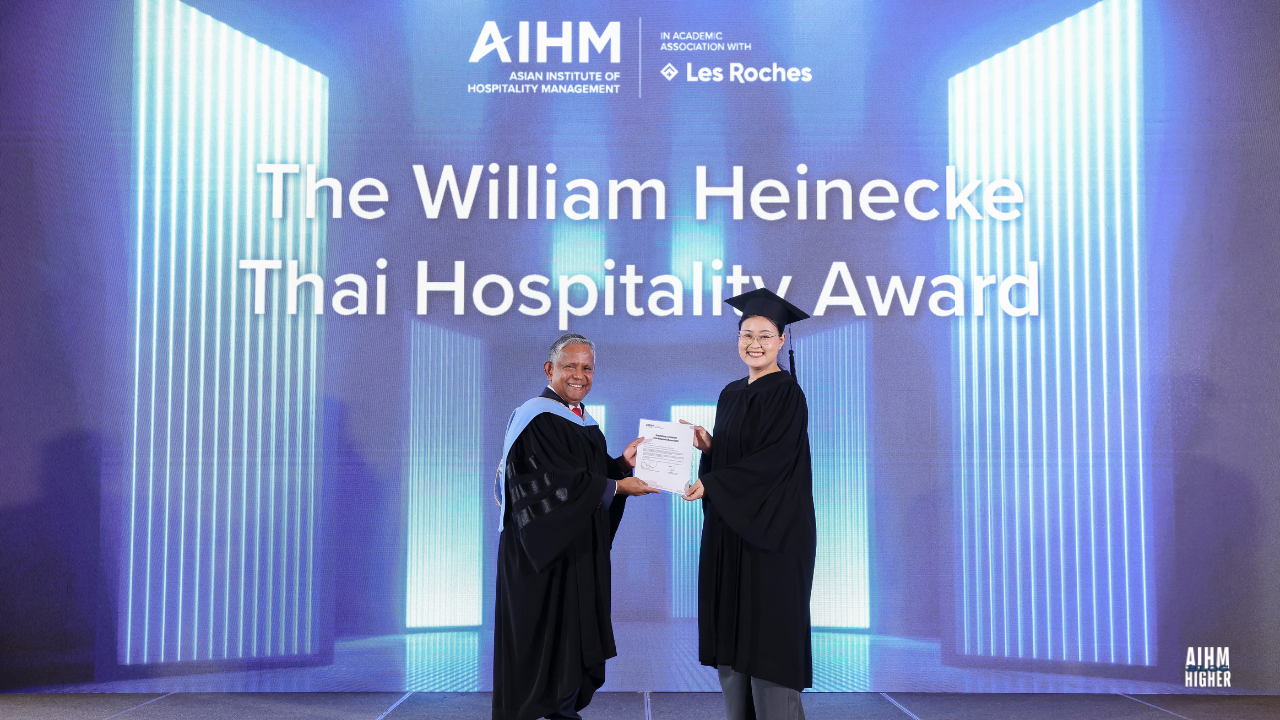 Patthamawan Dechasurakchon CIHA4 receives The William Heinecke Thai Hospitality Award from Mr. Dillip Rajakarier, CEO of Minor Hotel International and CEO of Minor Hotels during the AIHM Graduation Ceremony 2025. Both are smiling proudly on stage against a blue-lit backdrop featuring the award title.