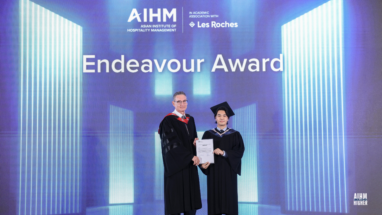 Joe Miller poses with Mr. Warren Stanworth, Academic Dean  after receiving the Endeavour Award during the AIHM Graduation Ceremony 2025. Both hold the certificate proudly in front of the glowing AIHM backdrop.