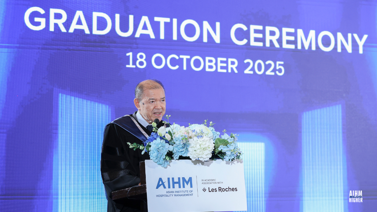 Khun Supachai Panitchpakdi, Chairman of the AIHM Board of Trustees in academic regalia delivers a speech at the AIHM Graduation Ceremony 2025, standing behind a podium decorated with blue and white flowers. The background screen displays the event date, “18 October 2025.”