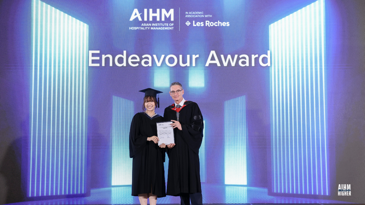 Kanpitcha Prapasnithikul joyfully receives the Endeavour Award from Mr. Warren Stanworth, Academic Dean during the AIHM Graduation Ceremony 2025. The blue and white illuminated stage backdrop highlights the event’s celebratory tone.