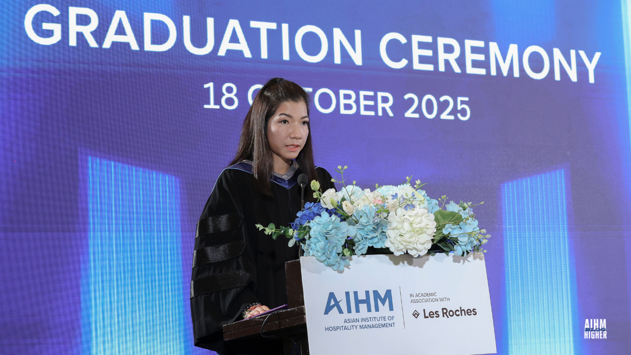 Dr. Pimporn Chawananth,Secretary to the Minister of Higher Education, Science, Research and Innovation in a graduation gown addresses the audience from a podium adorned with a blue and white floral arrangement during the AIHM Graduation Ceremony 2025. The event date is shown on the screen behind him.