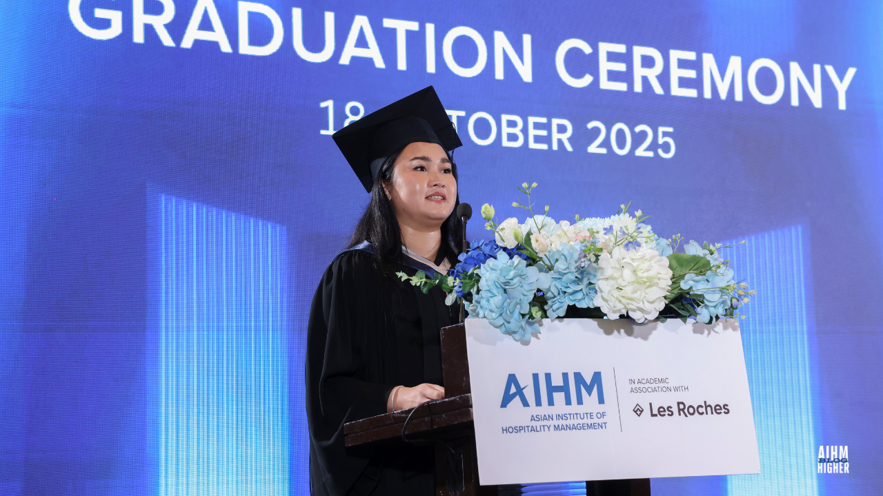 Sandy Sai Shan Davis BBA Cohort2 in academic regalia speaks confidently at the podium during the AIHM Graduation Ceremony 2025. The podium is adorned with a blue and white floral arrangement, and the screen behind displays the event title and date.