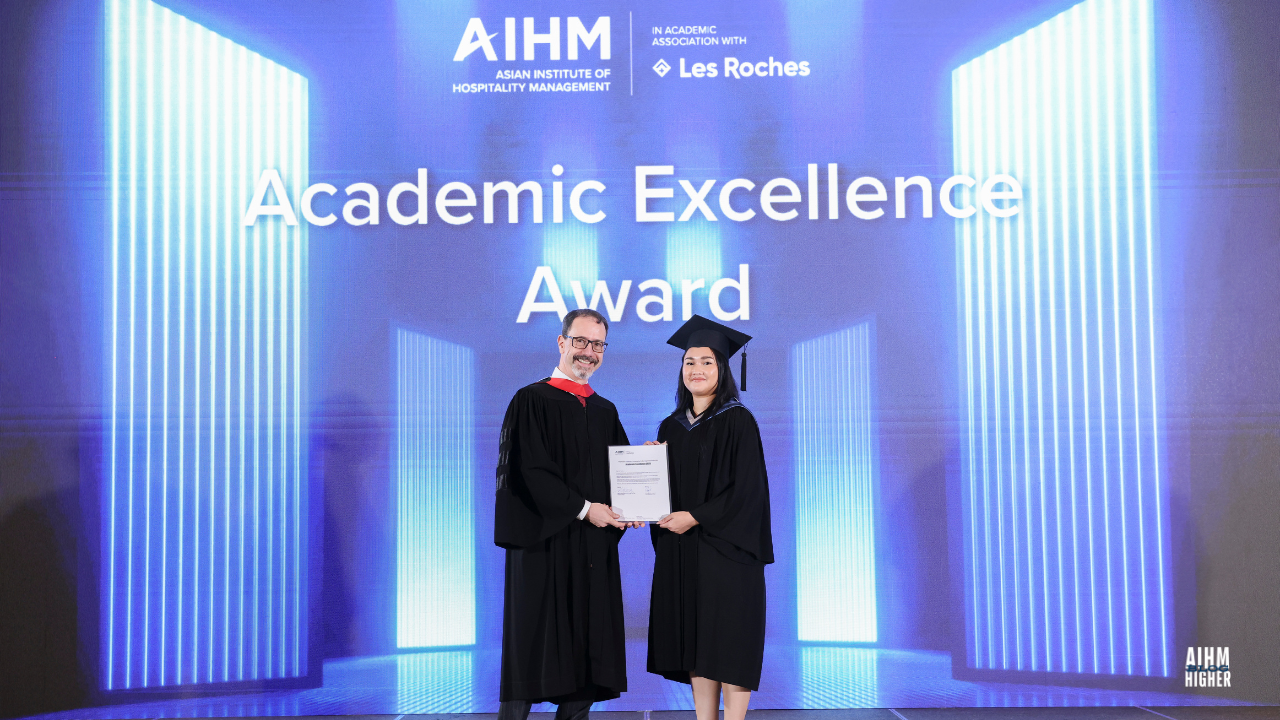 Sandy Sai Shan Davis smiling accepts the Academic Excellence Award from Mr. Chris Meylan, COO of AIHM during the AIHM Graduation Ceremony 2025. The bright blue stage background displays “Academic Excellence Award” along with the AIHM and Les Roches logos.