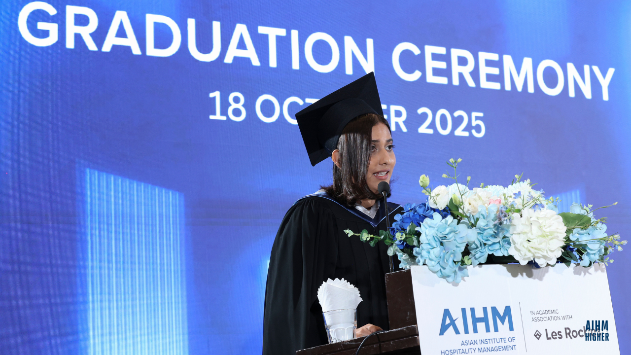 Maya Mariam BBA Cohort3 in cap and gown delivers a speech at the podium during the AIHM Graduation Ceremony 2025. The stage backdrop features the event title and date, “18 October 2025,” with blue and white floral decorations on the podium.