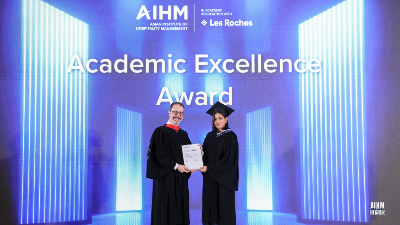 Maya Mariam in a AIHM gown and cap receives the Academic Excellence Award on stage during the AIHM Graduation Ceremony 2025. The award is presented by Mr. Chris Meylan, COO of AIHM in academic dress, with the AIHM and Les Roches logos displayed on a blue illuminated backdrop.
