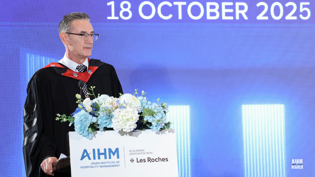 Warren Stanworth, Academic Dean in a graduation gown speaks from the podium adorned with blue and white flowers during the AIHM Graduation Ceremony 2025. The event date, “18 October 2025,” is displayed on the blue background behind him.
