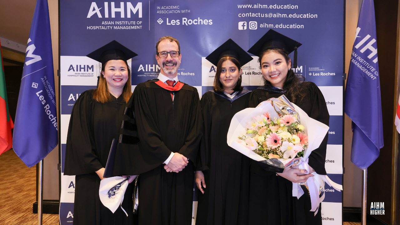 Three graduates and Chris Meylan, COO of AIHM in academic regalia smile together in front of the AIHM and Les Roches backdrop. One graduate holds a bouquet of flowers, marking the joyful celebration of AIHM Graduation 2025.